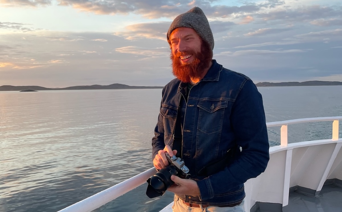 Quincy Morgan smiling with a red beard, grey knit cap, and jean jacket holding a Fuji camera aboard a ferry in Labrador at sunset at sea with the coastline in the background.